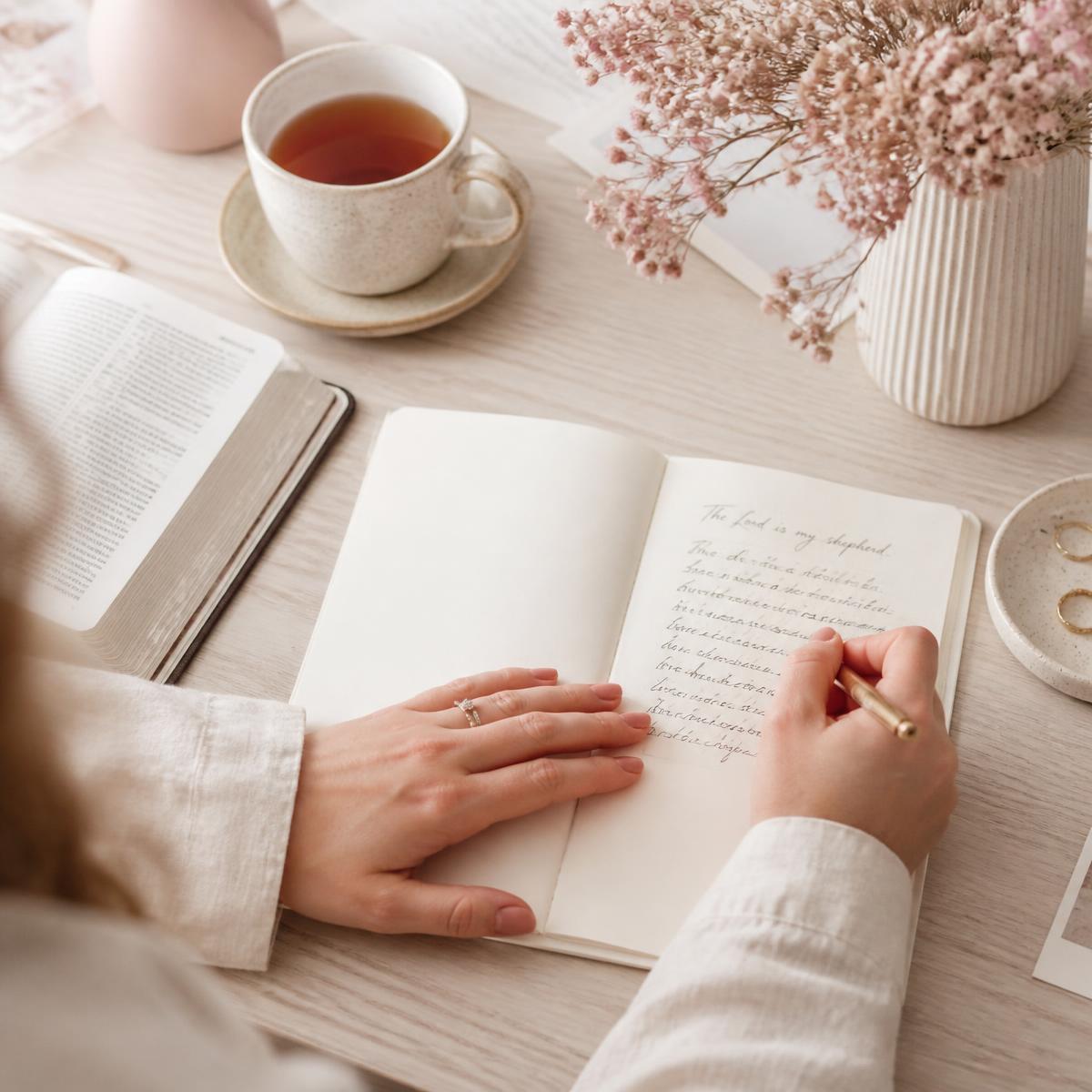Hands journalling beside an open Bible, tea and dried pink flowers